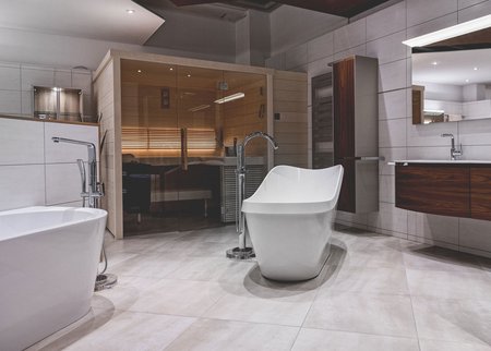 Moderne Badausstellung mit freistehender Badewanne, Glasdusche, großen Wandfliesen und einer integrierten Holzsauna.