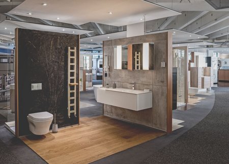 Ausstellungskabine mit dunkler Steinwand, hellem Holzfußboden, Wand-WC und Waschplatz mit beleuchtetem Spiegel.