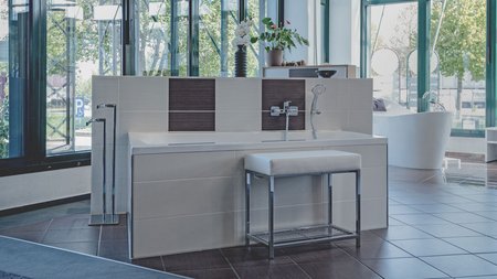 Badewanneninstallation mit Sitzbank, Wandarmatur, dekorativen Fliesenakzenten und großem Fensterbereich in einer Badausstellung.