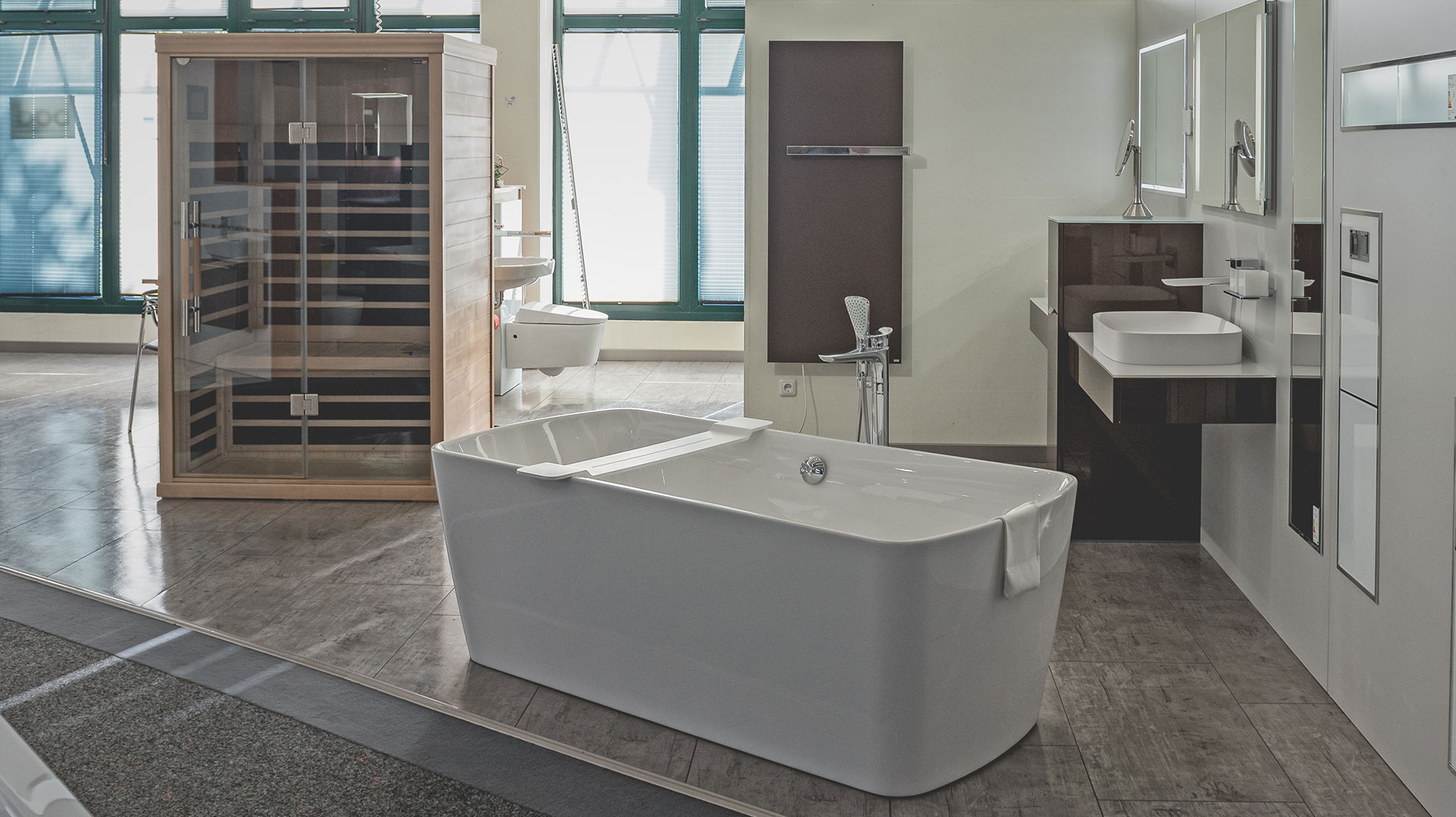 Großzügige Badausstellung mit weißer freistehender Badewanne, moderner Infrarotkabine aus Holz und Waschtischanlage mit großem Spiegel.